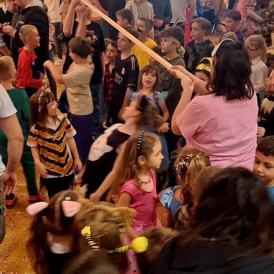 Eine Gruppe von Kindern und Erwachsenen ist drinnen versammelt. Einige Kinder tragen Kostüme, und eine Erwachsene hält einen langen Holzstab. Die Kinder sehen aufgeregt aus, und einige tragen Accessoires wie Stirnbänder und Halsketten.
