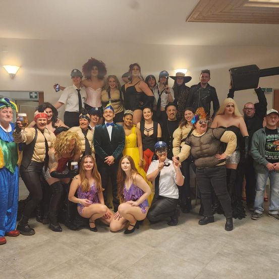 Eine Gruppe von Menschen in verschiedenen Kostümen posiert für ein Foto. Einige sitzen und andere stehen, halten Getränke und tragen Hüte und Brillen. Sie befinden sich wahrscheinlich auf einer Kostümparty.