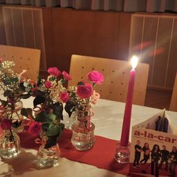 Ein Tisch mit weißer Tischdecke hat mehrere Glasvasen mit rosa Blumen und eine rote Kerze. Hinter dem Tisch stehen Stühle und eine Heizung.