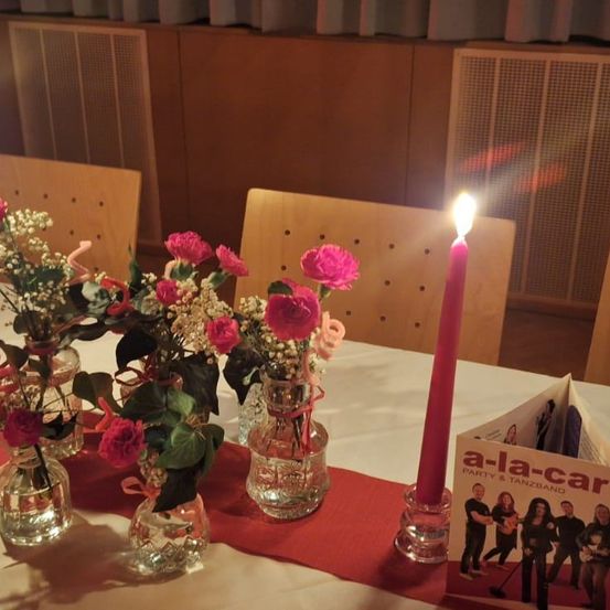 Ein Tisch mit weißer Tischdecke hat mehrere Glasvasen mit rosa Blumen und eine rote Kerze. Hinter dem Tisch stehen Stühle und eine Heizung.