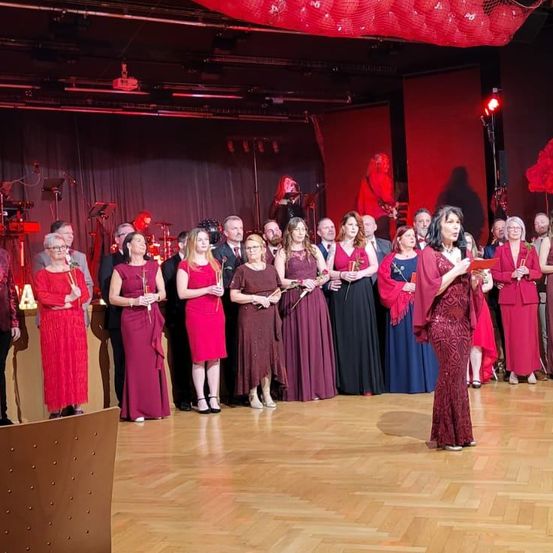 Eine Gruppe von Menschen steht auf einer Bühne, gekleidet in Rot und mit Rosen in den Händen. Eine Frau in der Mitte hält ein Buch und spricht in ein Mikrofon. Rote Lichter und Ballons schmücken die Bühne.
