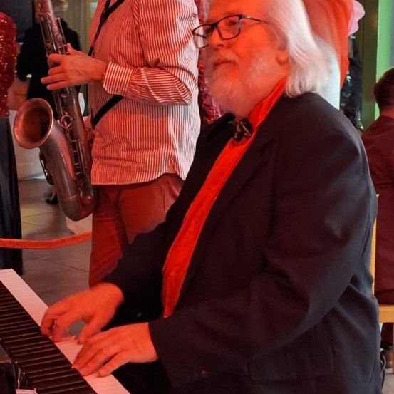 Ein älterer Mann spielt Klavier, während ein anderer Mann hinter ihm Saxophon spielt. Beide schauen konzentriert auf ihre Aufführung.