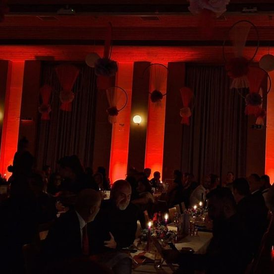 Ein schwach beleuchteter Bankettsaal mit roter Beleuchtung. Mehrere Menschen in Anzügen sitzen um Tische herum, einige mit brennenden Kerzen. Die Wände sind rot, und es gibt dekorative Aufhängungen.