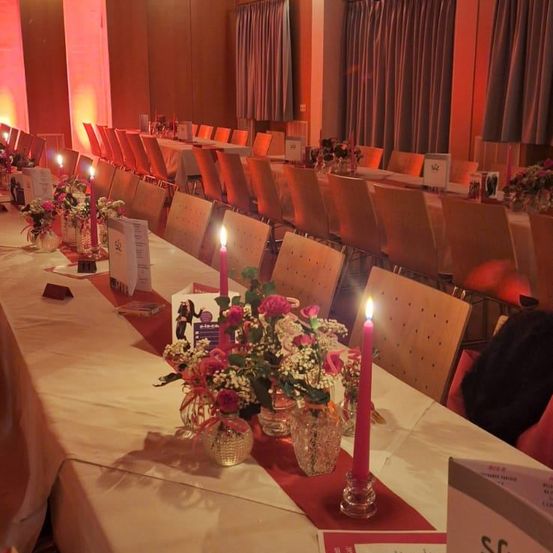 Ein langer Banketttisch für eine formelle Veranstaltung mit weißen Tischdecken, rosa Kerzen und Blumenarrangements. Der Raum ist mit roten Lichtern schwach beleuchtet.