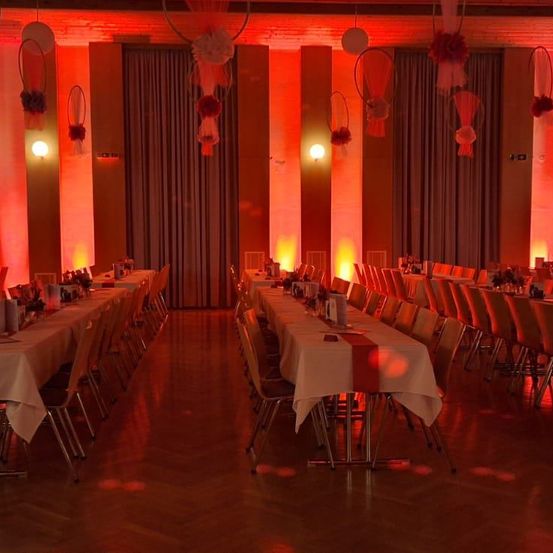 Ein leerer Bankettsaal ist mit roten Lichtern beleuchtet. Zwei lange Tische sind mit weißen Tischdecken und roten Tischläufern gedeckt. Dekorationen hängen von der Decke.