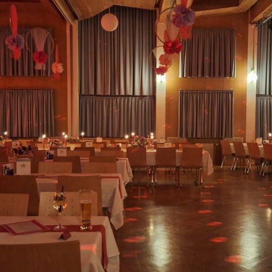 Ein Bankettsaal mit mehreren Tischen, jeweils mit Blumen und Kerzen. Dekorationen hängen von der Decke. Der Boden ist aus Holz.