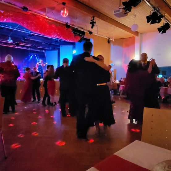 Mehrere Paare tanzen auf einer Ballsaalfläche mit roter Beleuchtung. Die Bühne hat eine Band, die mit einer roten Ballondekoration spielt. Die Leute sitzen an Tischen und schauen den Tanz.