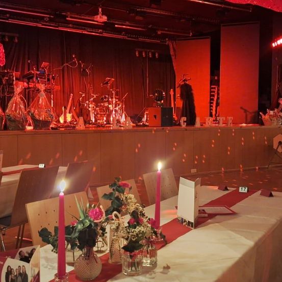 Eine Bühne mit einer Band-Aufstellung, einschließlich eines Schlagzeugs, Mikrofons und Instrumenten. Der Bereich ist schwach beleuchtet mit roter Beleuchtung. Es gibt Tische für ein Abendessen mit Blumen, Kerzen und Tellern gedeckt.