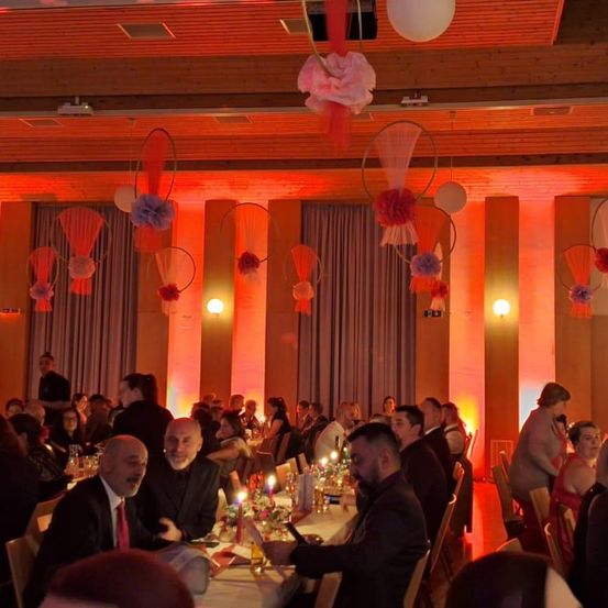 Eine Gruppe von Menschen sitzt an Tischen in einem Bankettsaal, mit Kerzen, Blumen und orangefarbenem Licht. Der Saal hat eine Holzdecke und Wände.