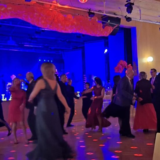 Menschen tanzen auf einer blau beleuchteten Tanzfläche mit Luftballons über ihnen, rote Lichter auf dem Boden. Männer und Frauen in formeller Kleidung bewegen sich zur Musik.