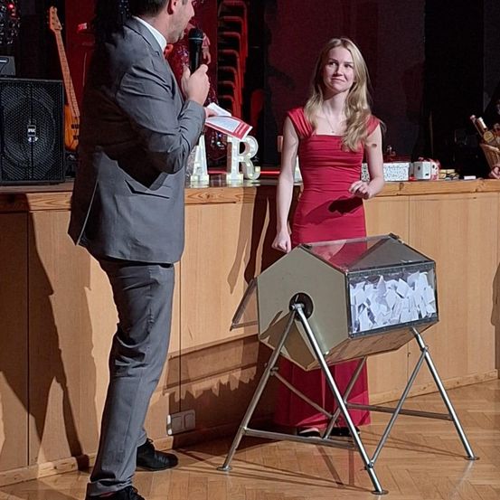 Ein Mann im Anzug hält ein Mikrofon und liest von einem Papier, er steht neben einer Frau in einem roten Kleid, mit einer Wahlurne zwischen ihnen auf der Bühne.