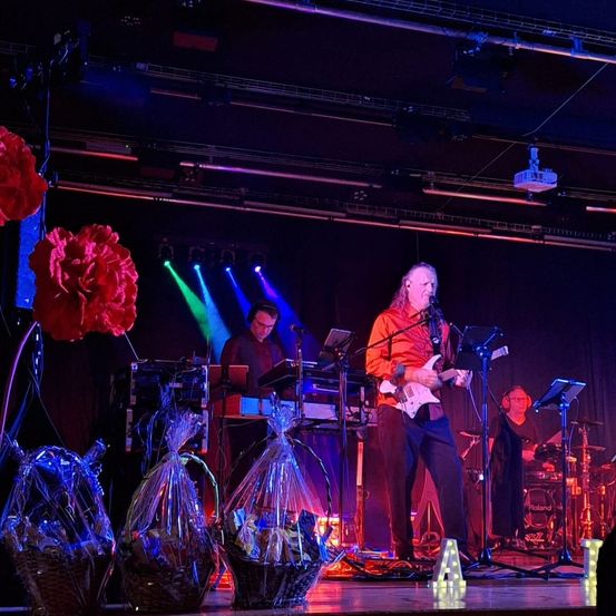Eine Band tritt auf der Bühne auf, beleuchtet von leuchtenden Farben. Ein Gitarrist steht mit einem Mikrofon, während ein Keyboarder und ein Schlagzeuger hinter ihm stehen. Die Bühne ist mit dekorativen Blumen und Geschenkkörben geschmückt.