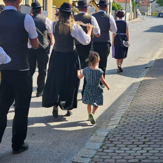 Eine Straßenszene mit einem Musikzug und einem kleinen Mädchen. Die Band spielt Instrumente, während das Mädchen entlanggeht. Eine Frau geht hinter ihnen.