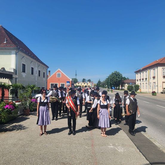 Eine Blaskapelle zieht mit Gebäuden, Bäumen und Blumen an der Straße entlang.