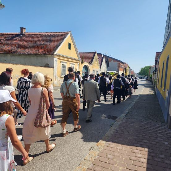 Eine Gruppe von Menschen in traditioneller Kleidung geht auf einer Straße. Die Straße wird von Gebäuden mit roten Dächern und gelben Wänden gesäumt. Sie scheinen an einer Prozession teilzunehmen.