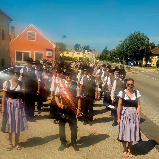 Eine Gruppe von Menschen in traditioneller bayerischer Kleidung, einschließlich Hüten und Schärpen, steht am Straßenrand während eines Festes. Dahinter sind Gebäude und Bäume zu sehen, und eine Straßenlaterne steht auf der rechten Seite.