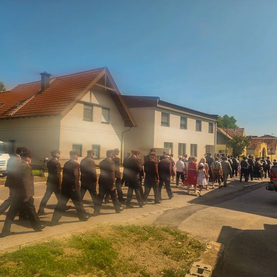Eine Gruppe von Menschen in formeller Kleidung geht vor einem Haus mit braunem Dach die Straße entlang. Dahinter gehen weitere Menschen. Der Bereich ist durch Tageslicht gut beleuchtet.
