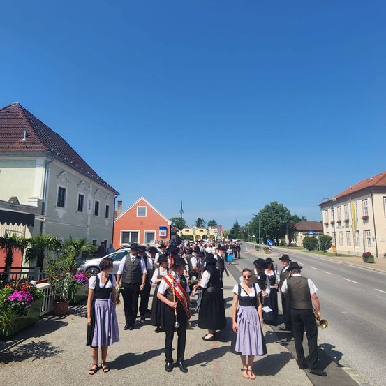 Eine Gruppe von Menschen in traditioneller Kleidung, die eine Straße hinuntermarschiert, mit einem klaren blauen Himmel darüber. Gebäude und Grünfläche säumen die Straße.