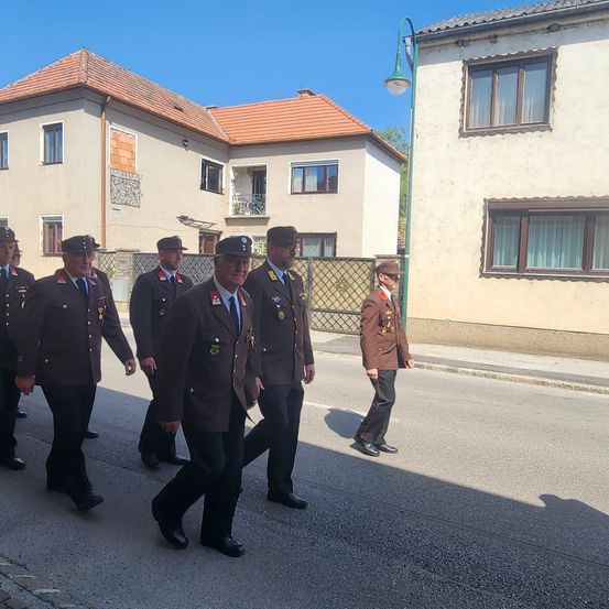 Eine Gruppe uniformierter Männer geht eine Straße entlang, mit Gebäuden auf beiden Seiten. Der Mann vorne führt die Gruppe.