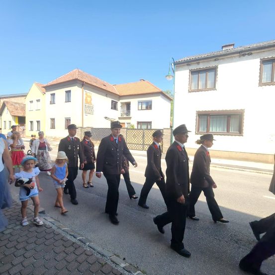 Mehrere Personen, einschließlich uniformierter Beamte und Zivilisten, gehen eine Straße entlang, die von Häusern gesäumt ist. Einige sind barfuß, und ein Kind hält ein Kuscheltier.