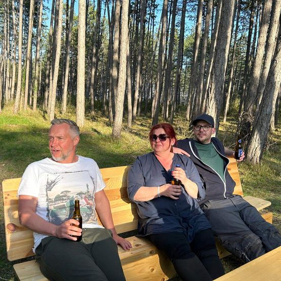 Drei Personen sitzen auf einer Holzbank im Wald. Sie halten Flaschen Bier in den Händen. Der Mann links trägt ein weißes T-Shirt. Die Frau in der Mitte hat eine Sonnenbrille auf. Der Mann rechts trägt ein grünes T-Shirt und eine Mütze.