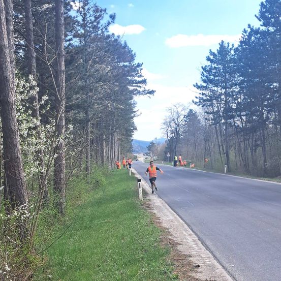 Ein Läufer in einem orangefarbenen Vest joggt auf einer ruhigen Straße, mehrere Personen in orangefarbenen Westen sind an der Seite. Die Straße ist von Bäumen gesäumt und Berge sind in der Ferne zu sehen.