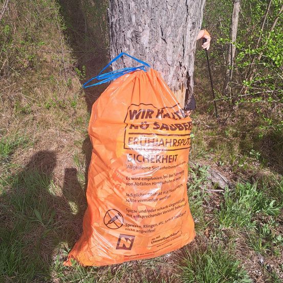 Ein orangefarbener Beutel mit 'Wir Halten Eicherheit' Text hängt an einem Baum im Wald. Er enthält Gegenstände wie Sprays und Flüssigkeiten.