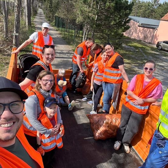 Eine Gruppe von Freiwilligen in orangenen Westen steht in einem Lastwagen mit Müllsäcken, lächelt und steht neben einem Haus und Bäumen.