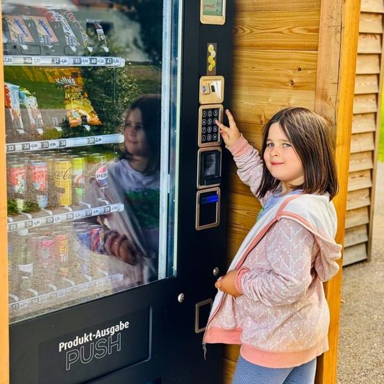 Ein junges Mädchen drückt einen Knopf an einem mit Snacks und Getränken gefüllten Automaten. Hinter der Scheibe sind ihr Spiegelbild und eine grüne Pflanze zu sehen.