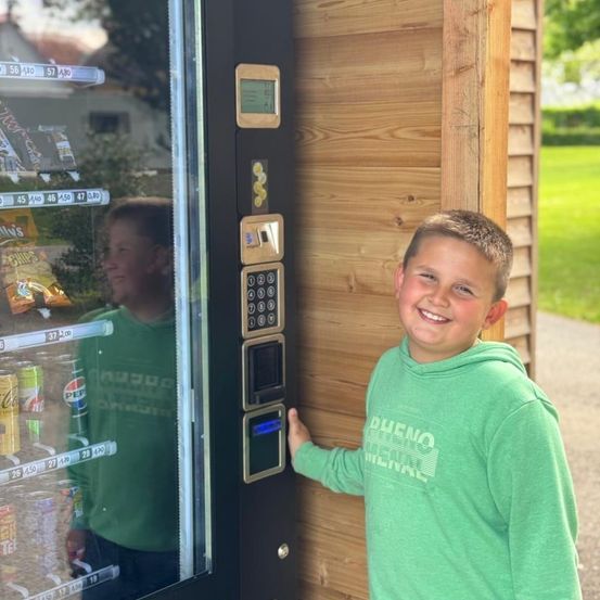 Ein junger Junge steht neben einem Automaten und lächelt. Der Automat hat verschiedene Snacks sichtbar und steht neben einer Holzwand.