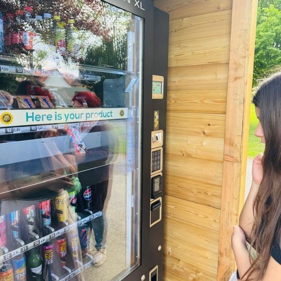 Eine Frau beobachtet eine andere Person, die ein Produkt aus einem Automaten holt. Der Automat zeigt eine Meldung mit der Aufschrift Hier ist Ihr Produkt.