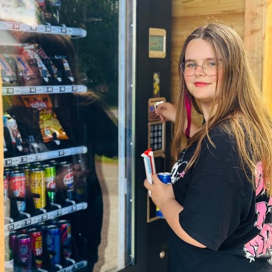 Eines Mädchen mit Brille steht bei einem Verkaufsautomaten und hält eine Dose. Der Automat hat Snacks und Getränke. Sie trägt ein schwarzes T-Shirt mit pinker Schrift.