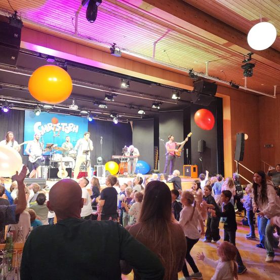 Eine Band spielt auf der Bühne mit bunten Luftballons und Lichtern. Kinder tanzen vor der Bühne, und ein Mann beobachtet vom Publikum aus.