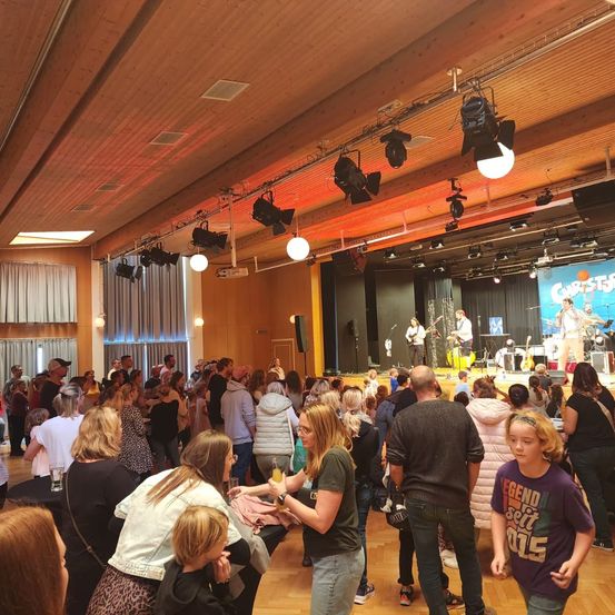 Eine Menschenmenge versammelt sich in einem großen Raum für eine Aufführung auf einer Bühne mit Instrumenten. Der Raum hat Holzböden, Wände und eine Decke mit Lichtern und Lautsprechern.