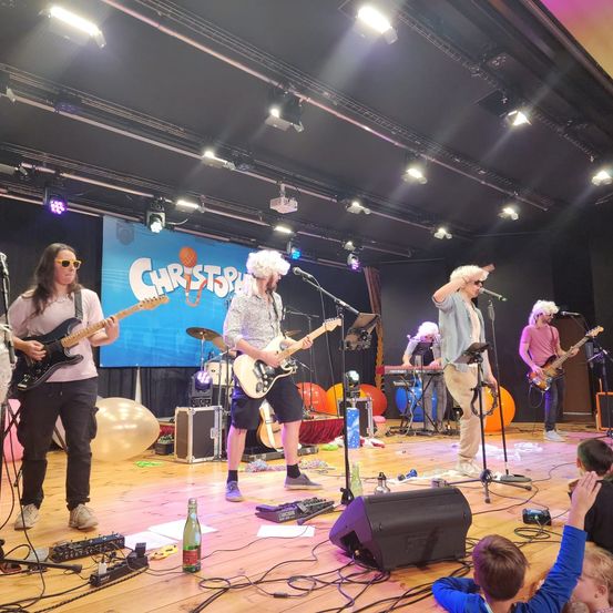 Eine Band spielt auf der Bühne mit Luftballons und einem farbenfrohen Hintergrund. Der Leadsänger singt in ein Mikrofon, während andere Gitarren und Keyboards spielen. Das Publikum schaut aufmerksam zu.