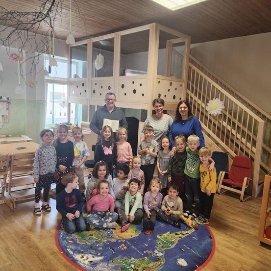 Eine Gruppe von Kindern und zwei Erwachsenen posiert für ein Foto in einem Raum mit einem Weltkartenteppich, Holzmöbeln und einer Treppe.