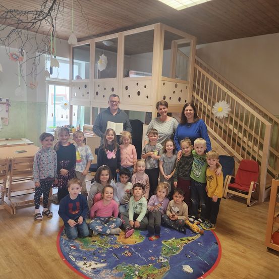 Eine Gruppe von Kindern und Erwachsenen in einem Klassenzimmer. Ein Mann und zwei Frauen stehen in der Mitte und halten ein Buch. Kinder sitzen auf einem Weltkartenteppich. Eine Treppe und ein Schreibtisch sind im Hintergrund.