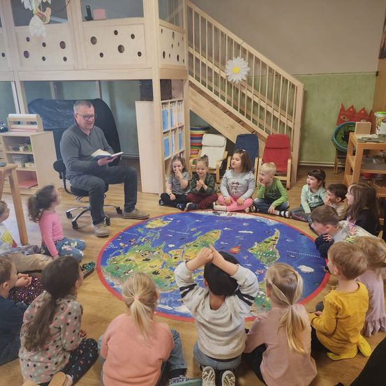 Ein Mann liest ein Buch vor einer Gruppe von Kindern vor, die im Kreis auf dem Boden sitzen, mit einer Weltkarte in der Mitte, in einem Spielzimmer mit Holzböden und Regalen.
