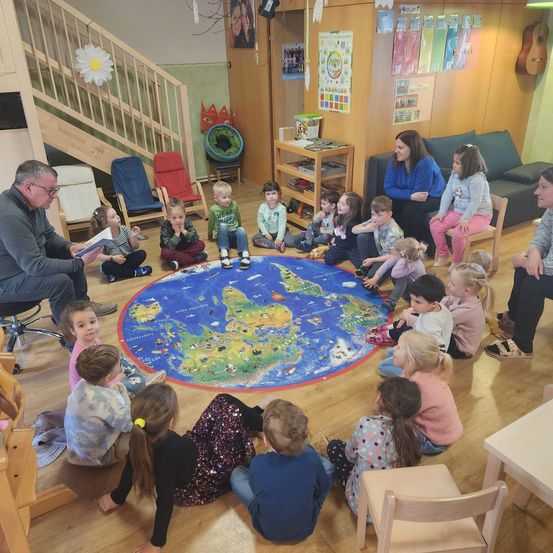 Eine Gruppe junger Kinder und Erwachsener sitzt in einem Kreis um eine Karte auf dem Boden. Ein Erwachsener liest ihnen ein Buch vor.