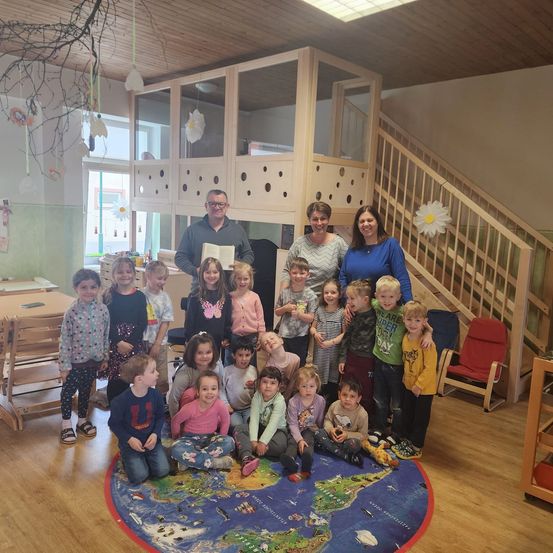 Eine Gruppe von Kindern und Erwachsenen steht um eine Weltkarte in einem Klassenzimmer. Ein Mann hält ein Zertifikat. Der Raum hat Holzböden, Regale und eine Treppe.