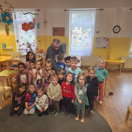 Eine Gruppe von Kindern und einem Erwachsenen in einem Klassenzimmer mit gelben Wänden und Holzböden, dekoriert mit Blumen und einer Uhr.