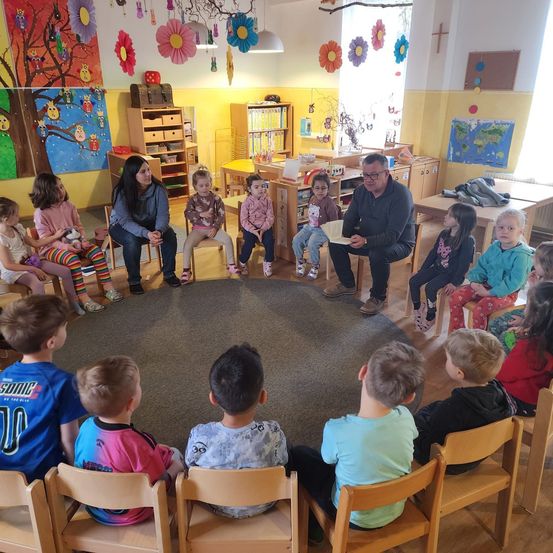Eine Gruppe von Kindern und zwei Erwachsenen sitzt in einem Kreis in einem Klassenzimmer. Der Lehrer liest aus einem Buch vor.