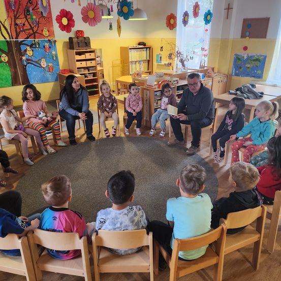Eine Gruppe von Kindern und zwei Erwachsenen sitzen in einem Klassenzimmer im Kreis. Der Lehrer liest ihnen ein Buch vor, während sie aufmerksam zuhören.