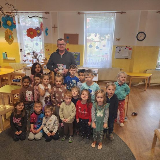 Ein Mann mit Brille steht in einem Klassenzimmer mit kleinen Kindern, lächelt und hält ein Kuscheltier. Der Raum hat gelbe Wände und Holzmöbel.