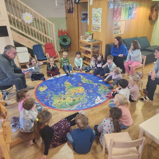 Eine Gruppe junger Kinder und Erwachsener sitzt um eine Weltkarte auf einem Holzboden. Ein Mann liest ihnen ein Buch vor, während andere zuhören. Der Raum hat Holzwände, eine Treppe und verschiedene Poster.