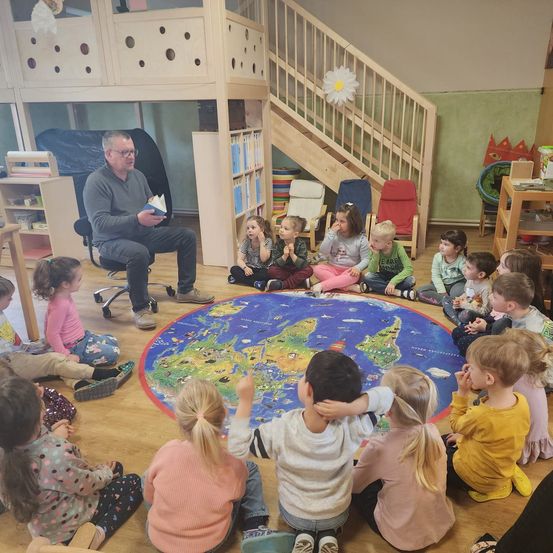 Ein Mann liest ein Buch vor einer Gruppe von Kindern vor, die in einem Kreis um eine Weltkarte auf dem Boden sitzen.