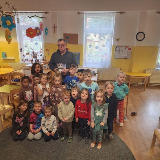 Eine Gruppe von Kindern in einem Klassenzimmer mit einem Mann, der ein Kuscheltier hält, posiert für ein Foto. Der Raum hat gelbe Wände, Fenster und eine Uhr an der Wand.