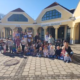 Eine Gruppe von Menschen, einschließlich Kindern, posiert für ein Foto vor dem Steinfeld Zentrum, einem Gebäude mit einem Café und einem Osterhasen-Maskottchen.