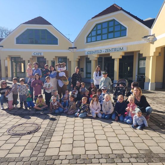 Eine Gruppe von Menschen, einschließlich Kindern, posiert für ein Foto vor dem Steinfeld Zentrum, einem Gebäude mit einem Café und einem Osterhasen-Maskottchen.