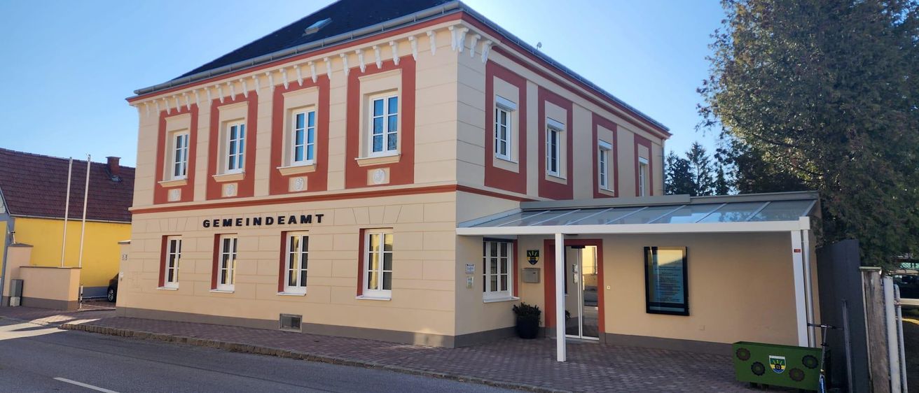 Ein zweistöckiges Gebäude mit einem Schieferdach steht an einer Straße. Es ist als 'Gemeindeamt', ein Rathaus, gekennzeichnet. Das Gebäude hat einen verglasten Eingang und wird von einem gepflasterten Bürgersteig flankiert. Ein Topf mit Pflanzen steht vor dem Eingang, und ein Mülleimer befindet sich auf der rechten Seite.